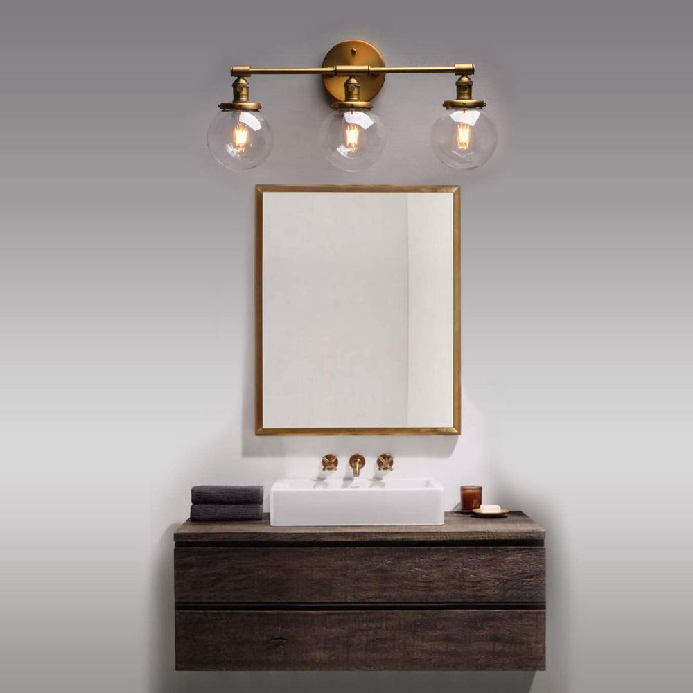 Three Light Bathroom Vanity Lighting with Clear Glass Globes with antique brushed gold hardware shown over a bathroom vanity